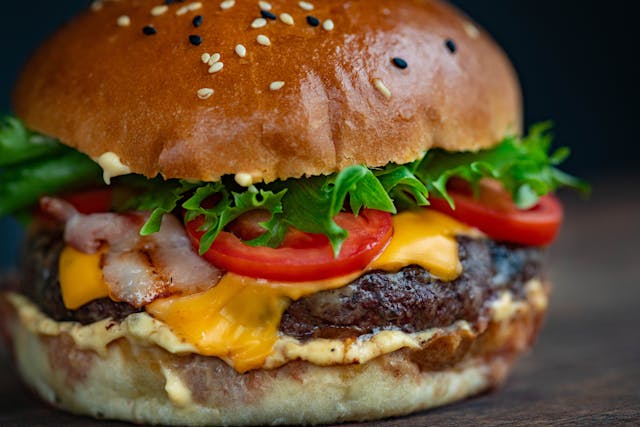 Close-up of a cheeseburger with a sesame seed bun, leafy lettuce, tomato slices, crispy bacon, melted cheddar cheese, and a beef patty, perfect for casual dining, with sauce spread on the bottom bun.