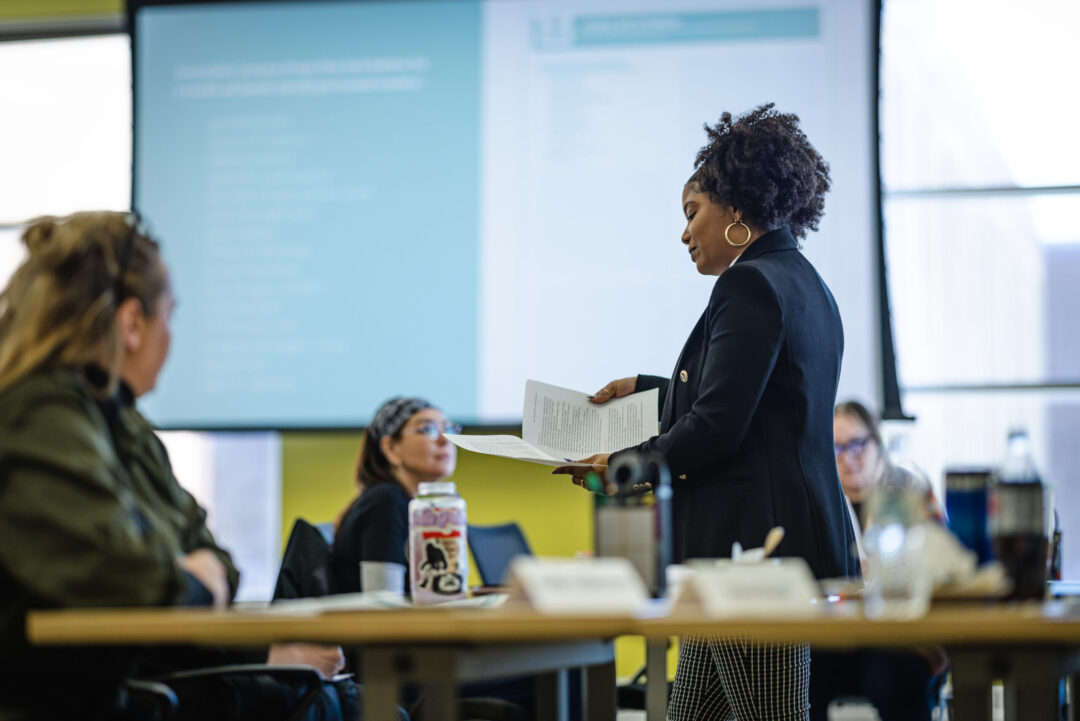 A woman stands and holds papers while speaking to a group seated around a table in a meeting room, leading a counseling session. A large screen with blurred text is displayed in the background.