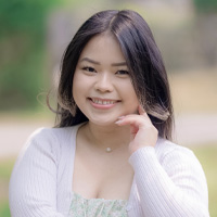 A young woman with long dark hair smiles gently, resting her hand on her chin. She wears a light-colored top and stands outside with a soft, blurred green and yellow background.