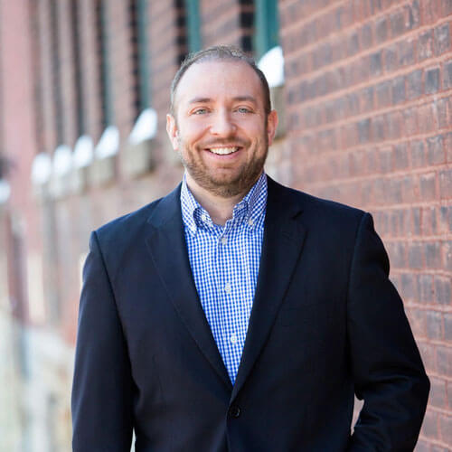 A man in a dark blazer and blue checkered shirt smiles while standing outside in front of a brick wall, reflecting on past events and activities.