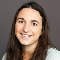 A woman with long brown hair and a light-colored top is smiling at the camera against a plain gray background.