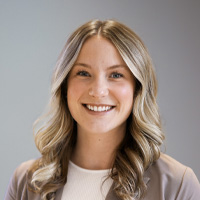 A woman with long, wavy blonde hair wearing a light brown blazer and white top, smiling at the camera against a plain, gray background.