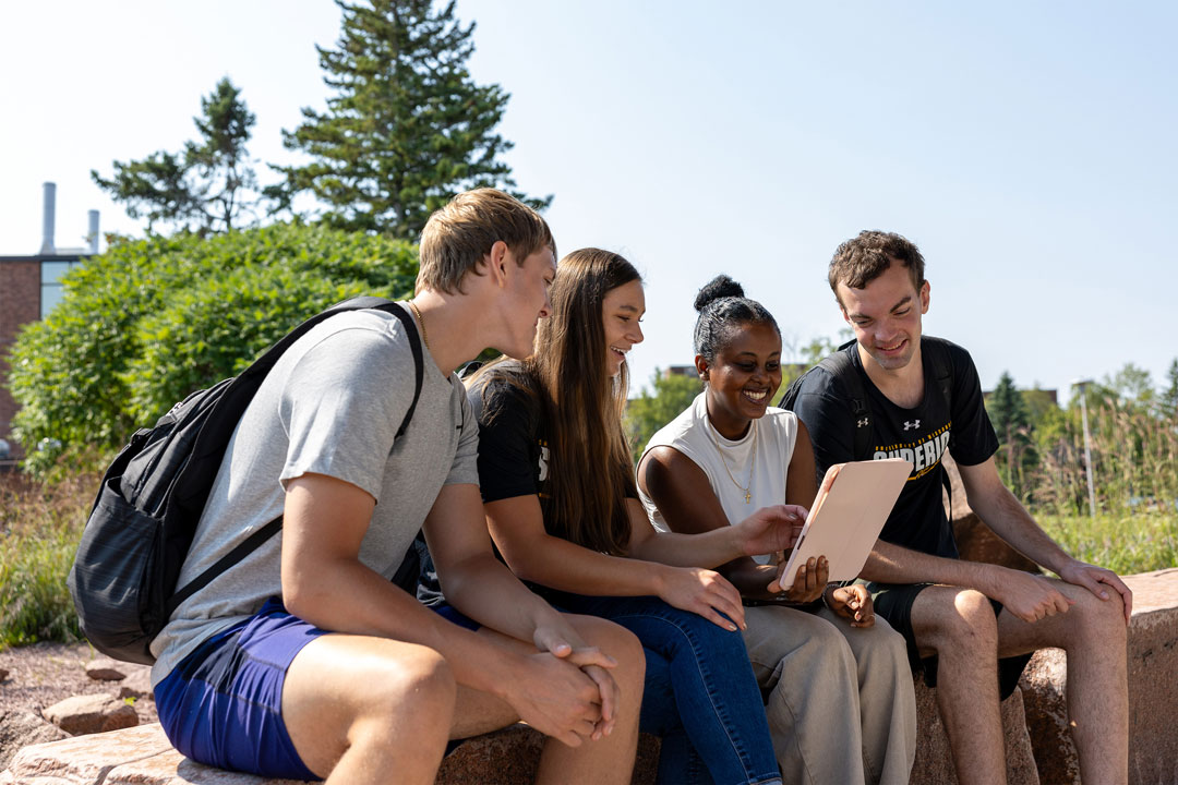 Four college students sit on a stone bench outdoors, smiling and looking at a tablet together on a sunny day, possibly reviewing a freshman application amid trees and greenery in the background.