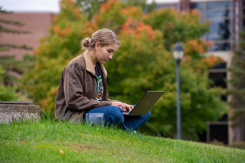 A student sits on a grassy slope outdoors, typing on a laptop. Trees with fall foliage and a University of Wisconsin-Superior building are blurred in the background. The person wears a brown jacket and has light brown hair pulled back.