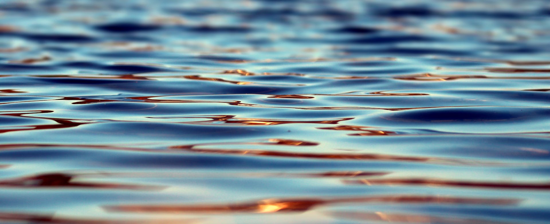Close-up view of gently rippling water near a Lake Superior research vessel for scientists, reflecting blue and golden tones from the sunlight and creating a smooth, abstract pattern across the surface.