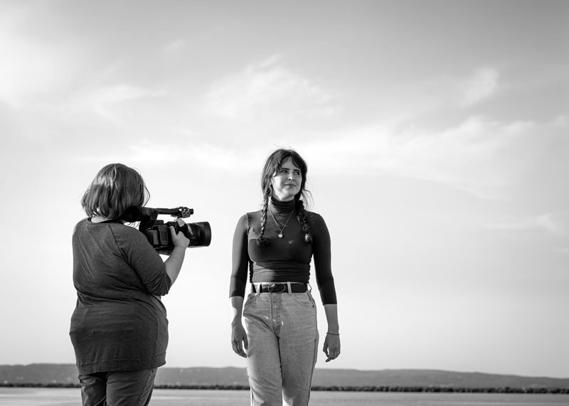 A person films another woman outdoors, capturing multimedia journalism against cloudy hills.