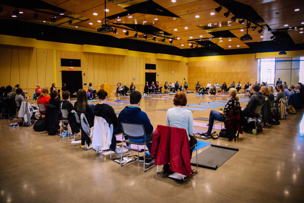 A large group of people sit in a wide circle on chairs and yoga mats in a spacious, well-lit room at the Pruitt Center for Mindfulness and Well-Being, attentively participating in a group event or meeting.