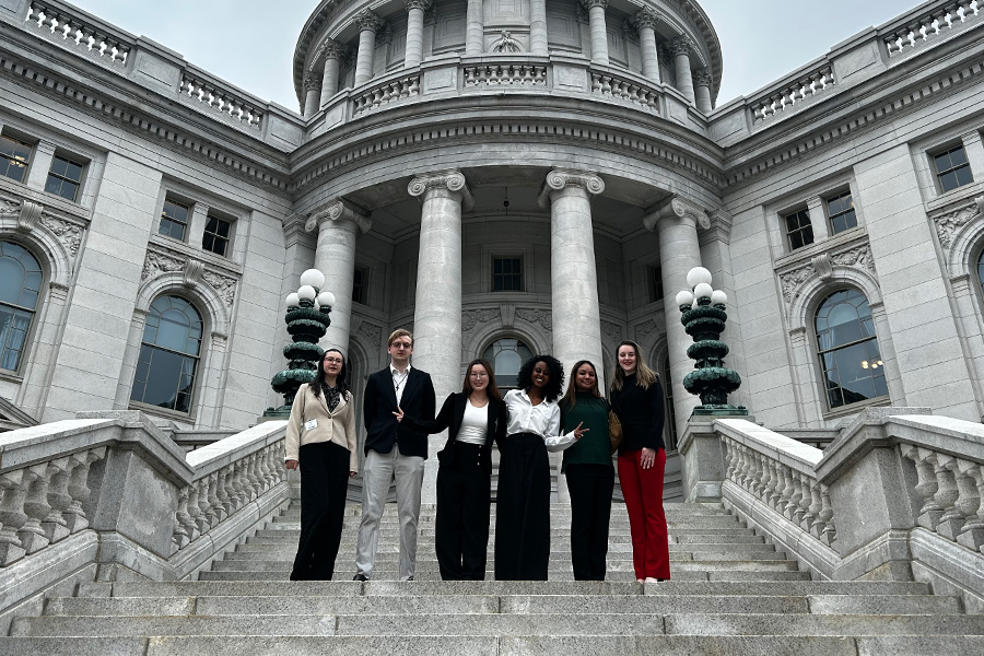 Six people stand on the steps of a grand, white stone building with columns and ornate architecture, posing and smiling for the camera. The sky is overcast.