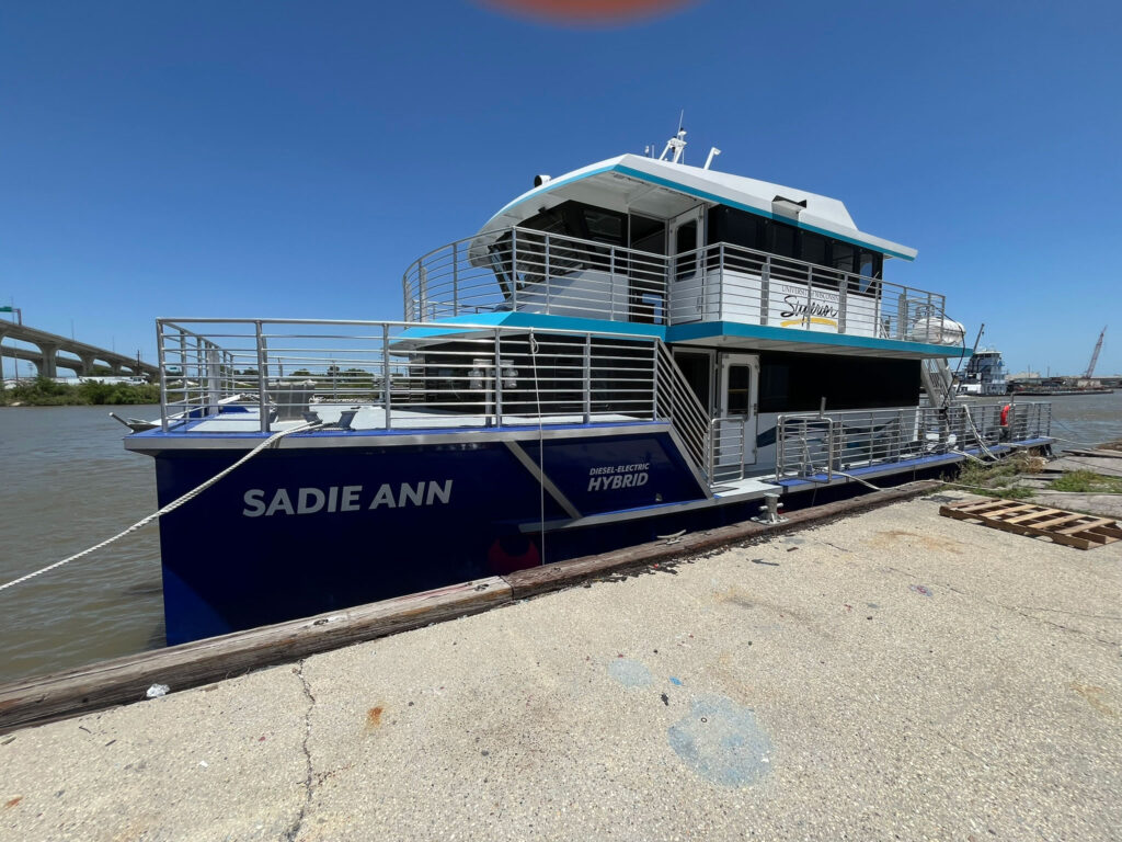 A blue and white boat named SADIE ANN is docked by a concrete pier on a sunny day, with railings, upper and lower decks, and water in the background. The words DIESEL ELECTRIC HYBRID are visible on the side, hinting about its eco-friendly technology.
