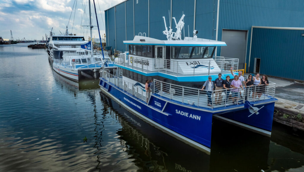 A group of people stands on the deck of the Sadie Ann research vessel in Lake Superior, docked beside another boat in a calm harbor with an industrial building visible in the background.