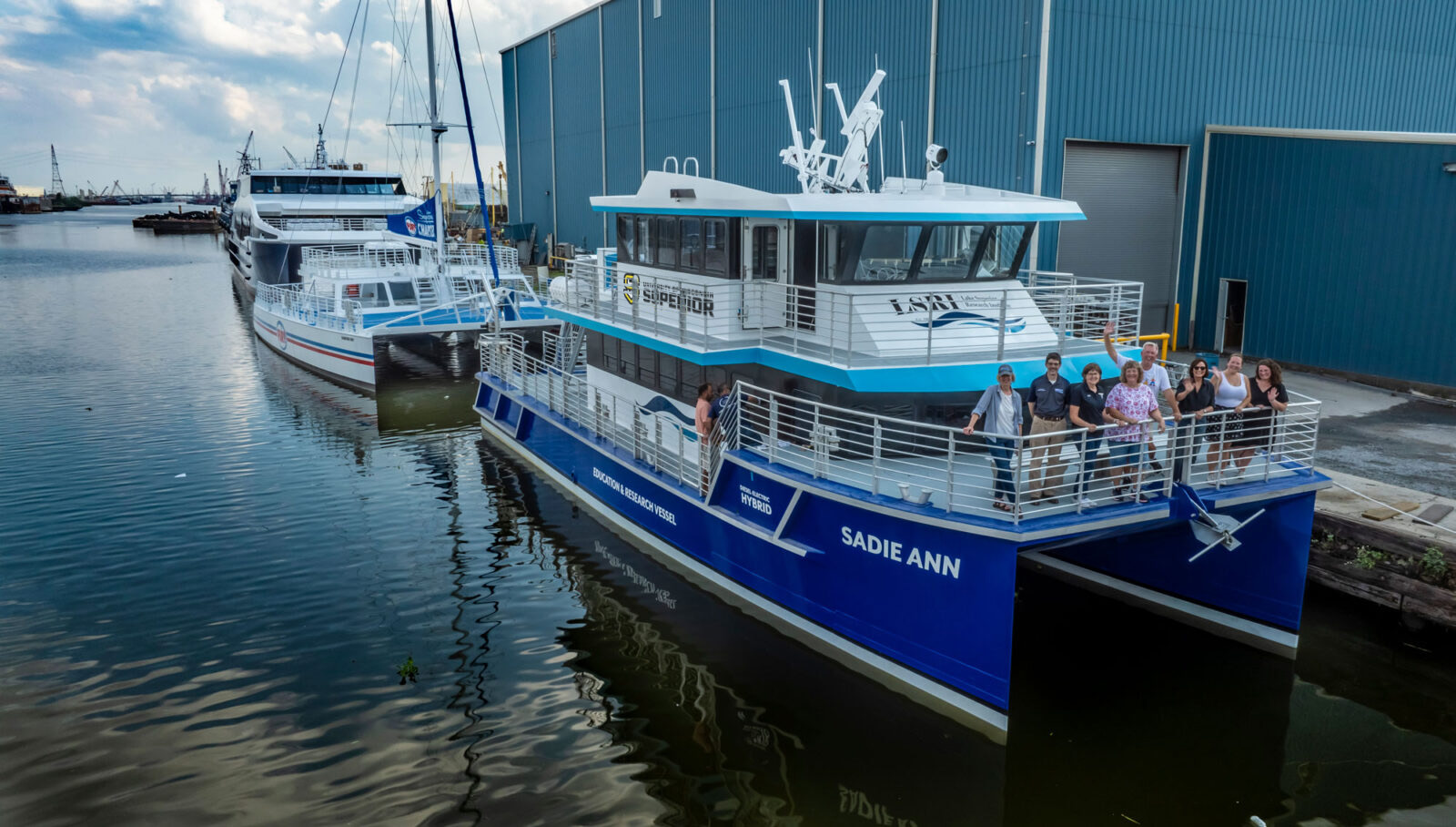 A group of people stands on the deck of the Sadie Ann research vessel in Lake Superior, docked beside another boat in a calm harbor..
