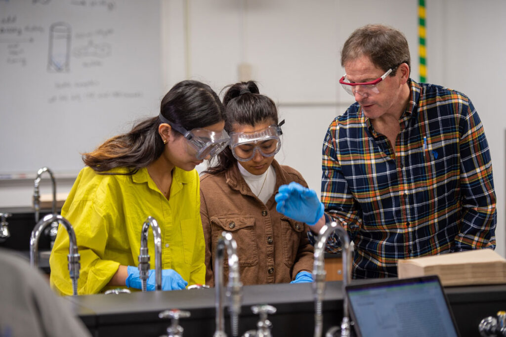 A teacher in a plaid shirt helps two students—future alumni—in goggles and gloves with a science experiment at a lab table. A whiteboard with diagrams is visible in the background.