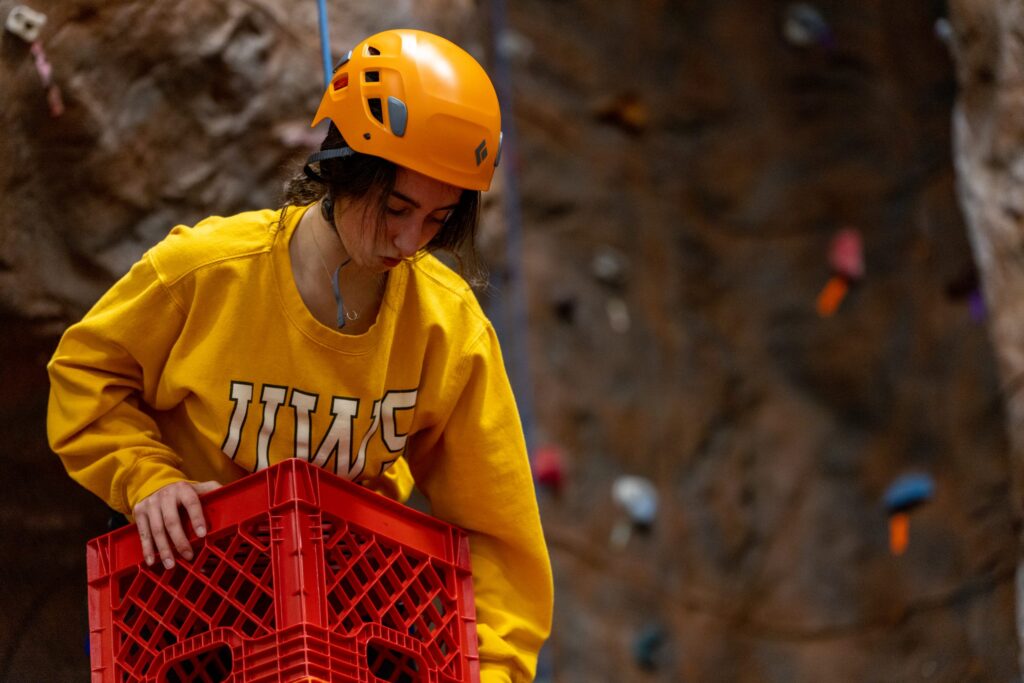 A person wearing a yellow helmet and yellow sweatshirt stands on stacked red crates in an indoor climbing gym, with a rocky climbing wall and colorful holds in the background.