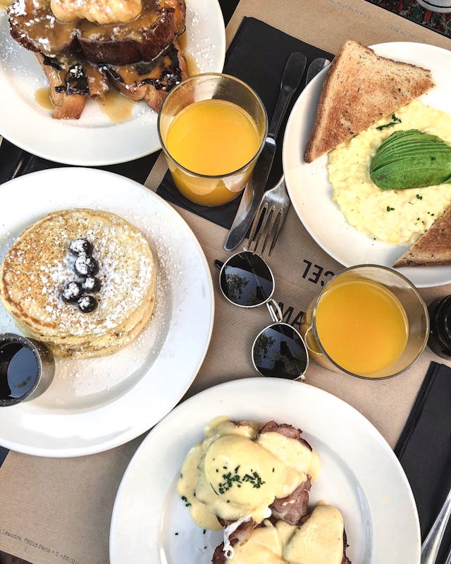 Four plates of breakfast food are set for dining: pancakes with blueberries, French toast with apples, scrambled eggs with toast and avocado, eggs Benedict—plus orange juice, coffee, and a pair of sunglasses on the table.