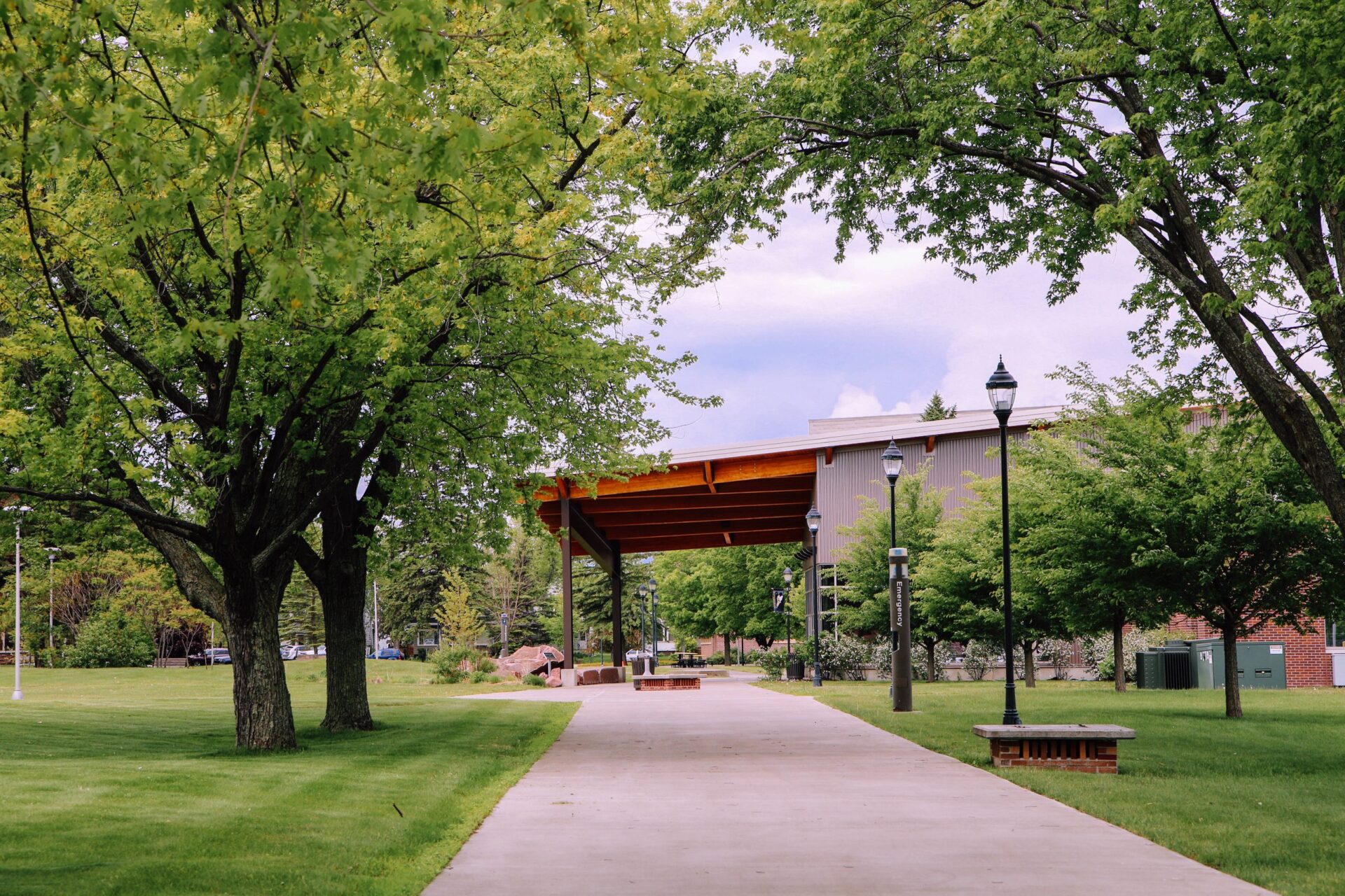 A paved walkway lined with green trees leads to a wooden pavilion in a park-like setting, with benches and lampposts along the path and a building visible in the background.