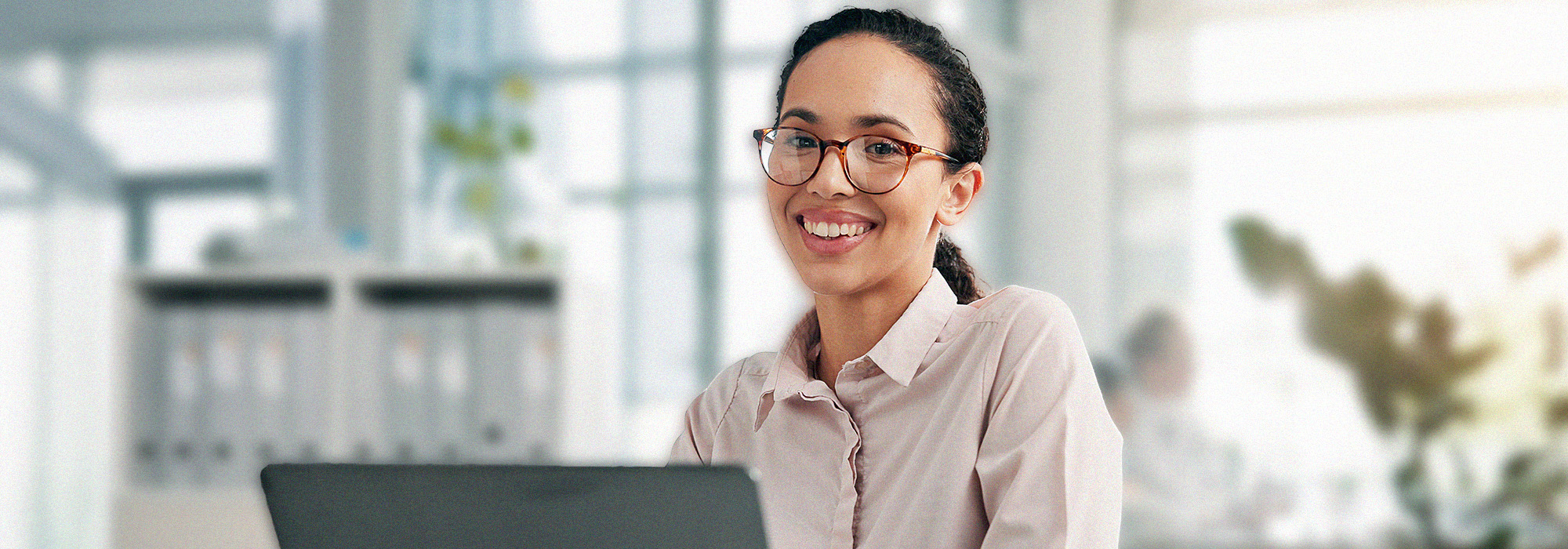 A woman with glasses and a light pink blouse smiles while sitting at a desk with a laptop in a bright, modern office, reflecting the professionalism of a Master of Social Work UW-Superior student.