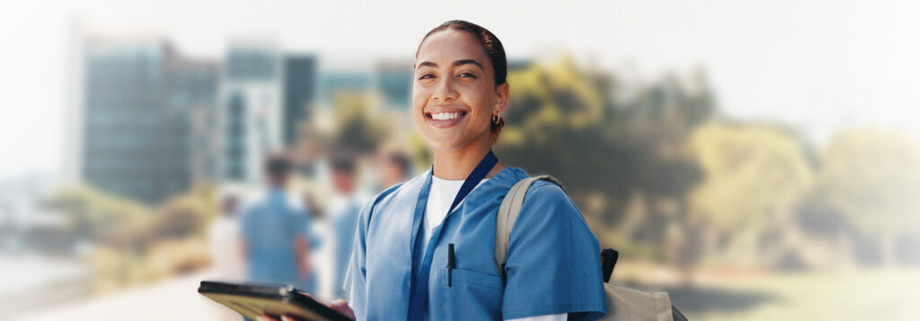 A smiling person wearing blue scrubs and a backpack holds a tablet outdoors, with blurred buildings and trees in the background.