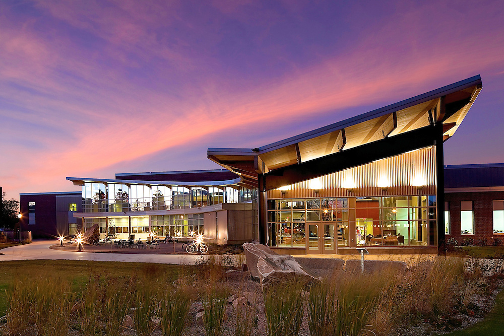 A modern building with large glass windows is illuminated at dusk, with warm lights inside and a vibrant purple and orange sky overhead. Grasses and rocks decorate the landscaped foreground.