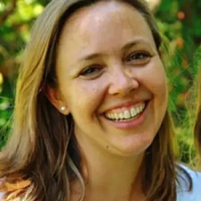 A woman with light brown hair smiles warmly at the camera, wearing a light-colored top. She is outdoors with a blurred green background, perhaps reminiscing about past events and activities that bring her joy.